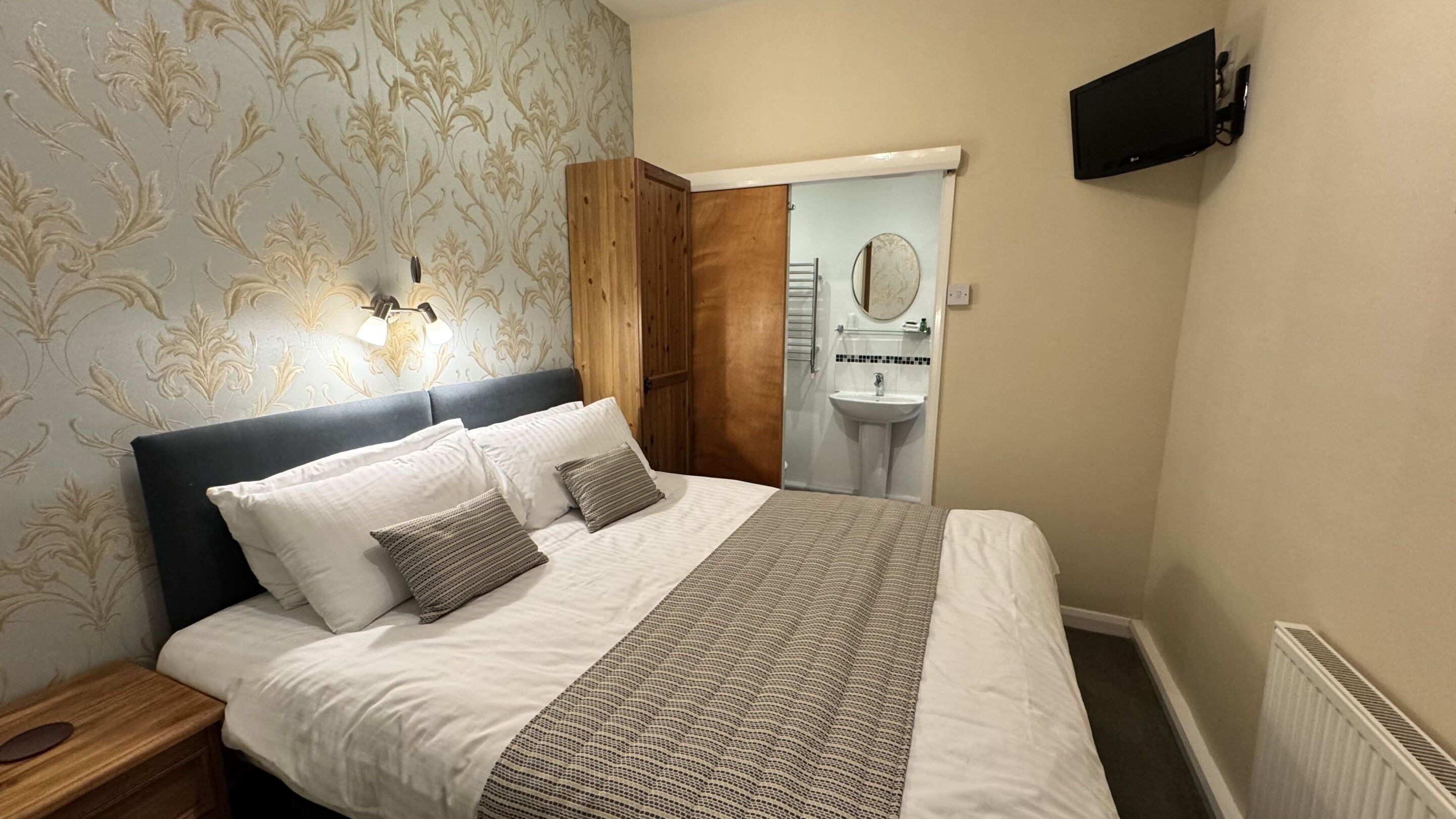 A bedroom with a bed, nightstand, wall-mounted TV, and a bathroom visible through an open door.