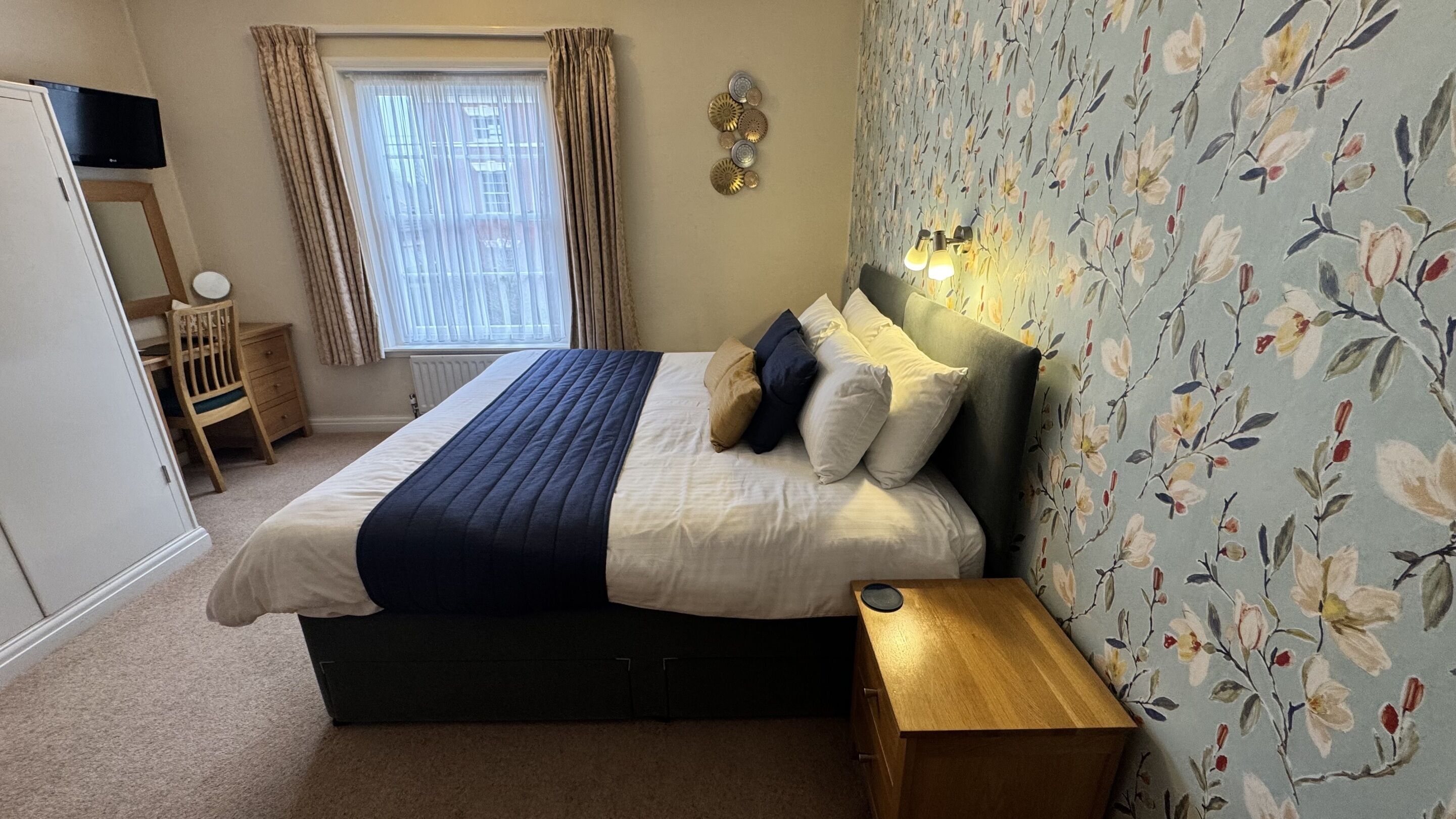A bedroom with a bed, a bedside table, a TV, a window with curtains, and a floral wallpaper.