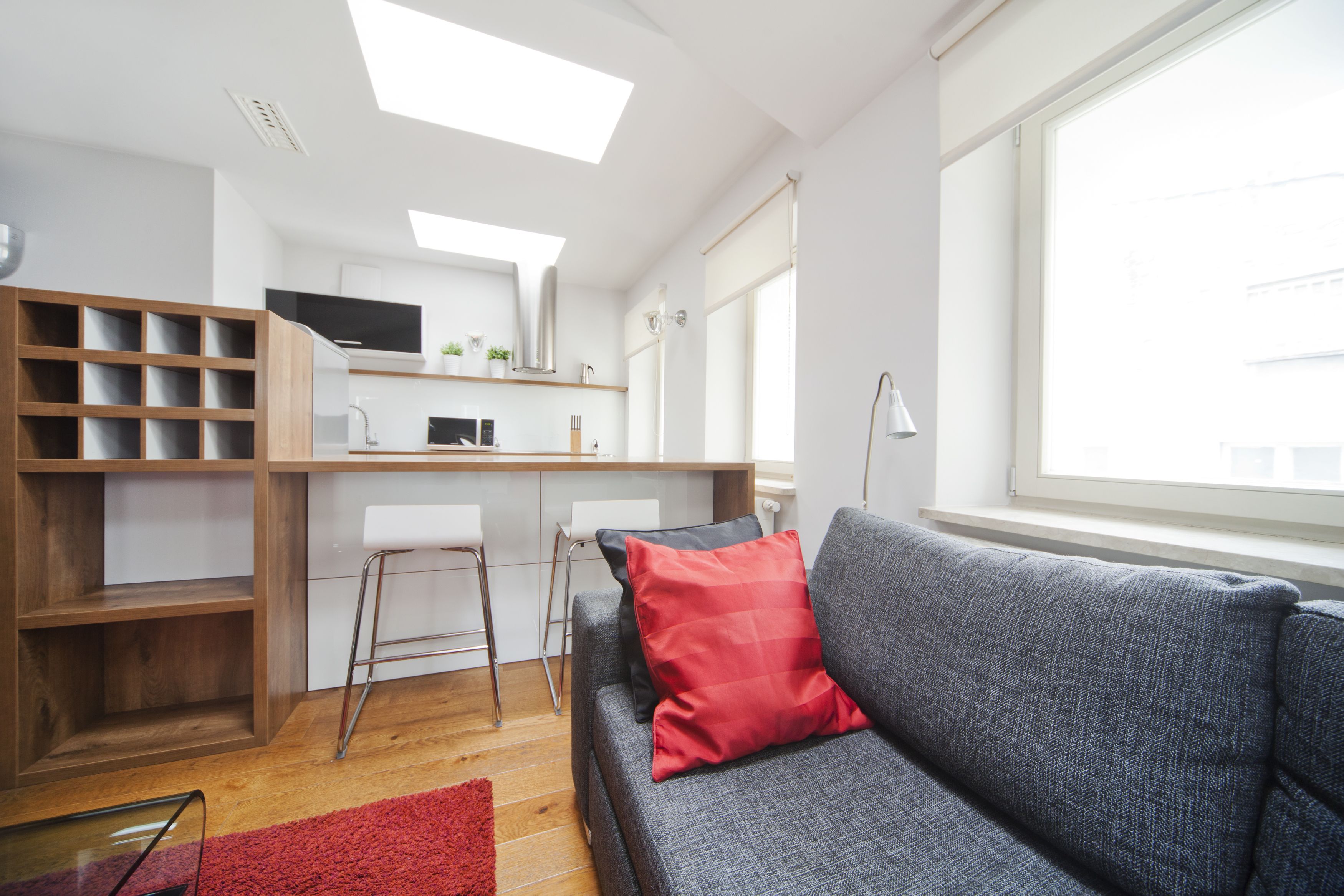 Una sala moderna con un sofá gris, una almohada roja, una estantería de madera y una zona de cocina con armarios blancos y un televisor.