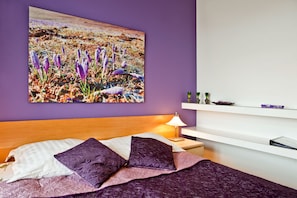 A bedroom with a bed, purple bedding, a nightstand, a lamp, a white shelf with decorative items, and a wall-mounted painting of flowers. - 4Seasons Apartments Cracow (Kraków)