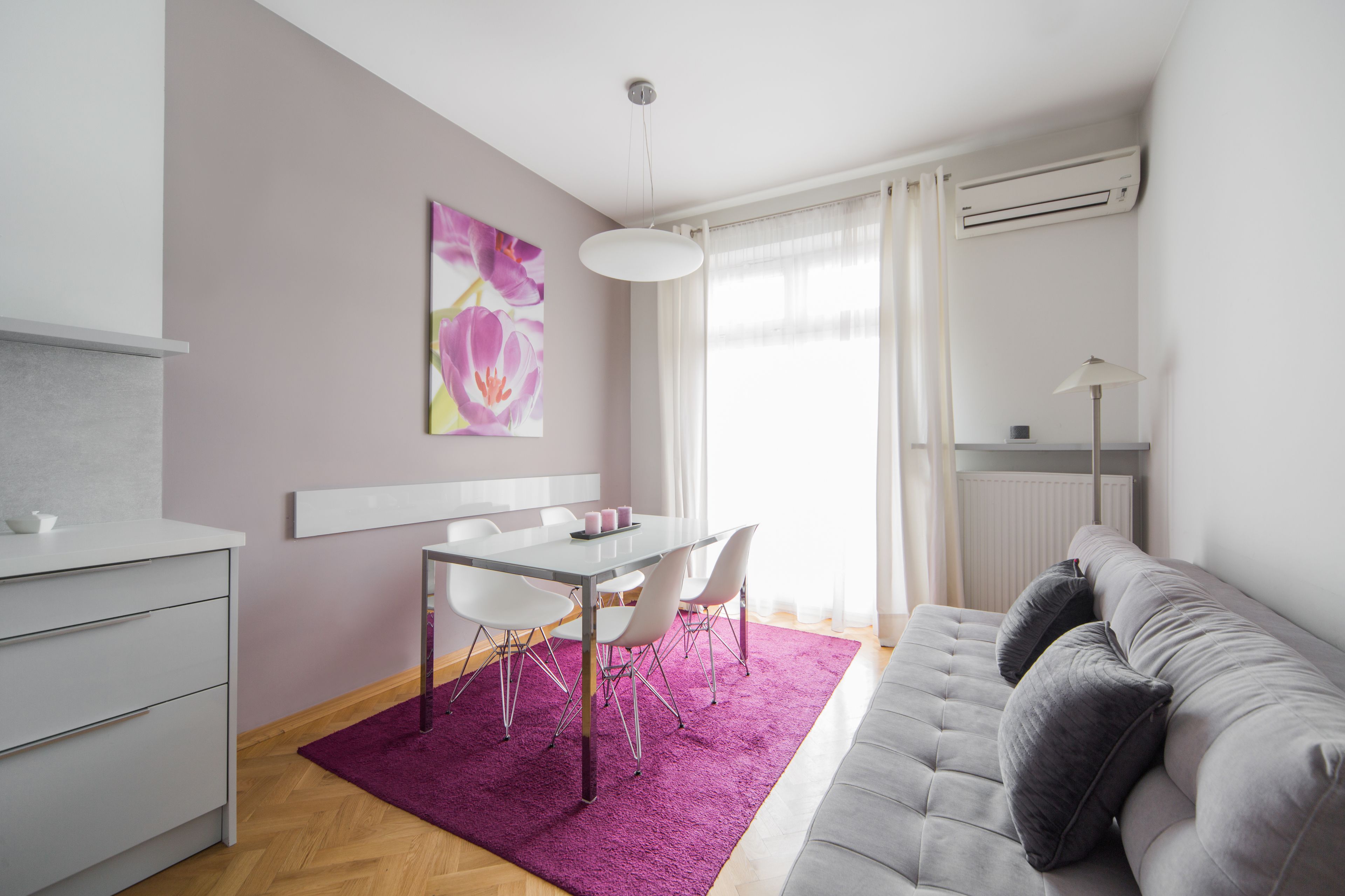 A modern living room with a grey sofa, a dining table with chairs, a purple rug, and a painting on the wall.