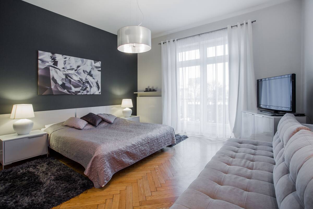 A modern bedroom with a bed, a sofa, a TV, and a large window with curtains.