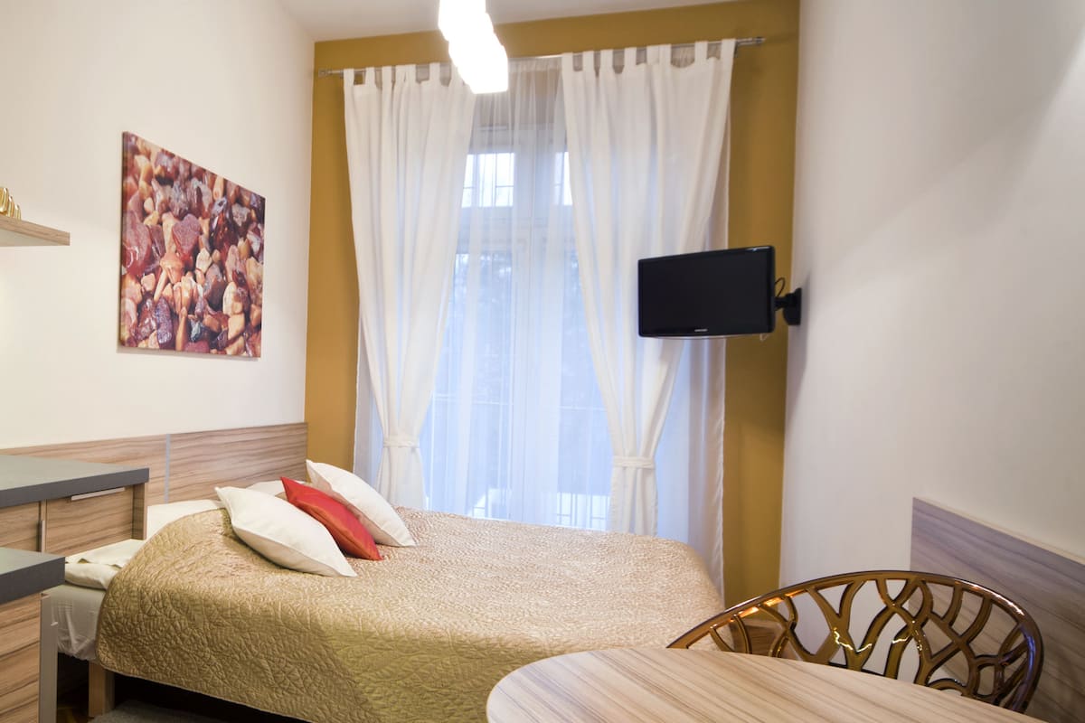 A bedroom with a bed, a desk, a chair, a TV mounted on the wall, and a window with curtains.