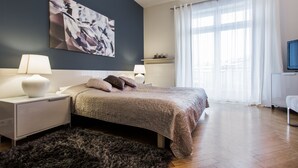 Un dormitorio moderno con cama, mesitas de noche, un cuadro abstracto grande, una ventana con cortinas translúcidas y un televisor sobre un mueble.
