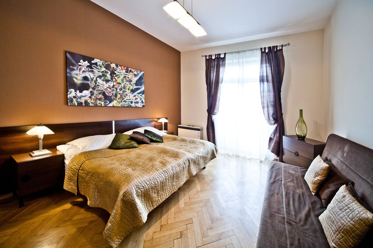 A hotel room with a large bed, a bedside table with a lamp, a nightstand, a dresser, a window with curtains, and a painting on the wall.