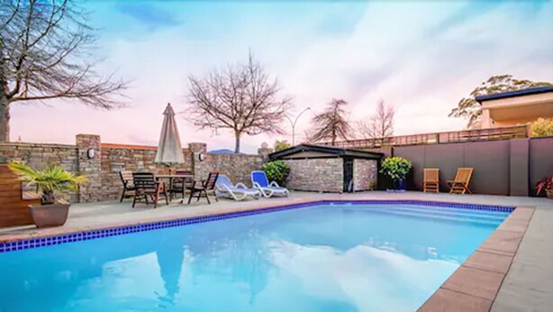 A swimming pool with lounge chairs and a table, surrounded by a stone wall and trees. - Tuscany Villas Rotorua (Rotorua)