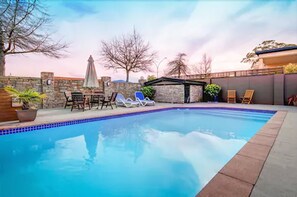 Piscina com espreguiçadeiras e mesa, rodeada por um muro de pedra e árvores.