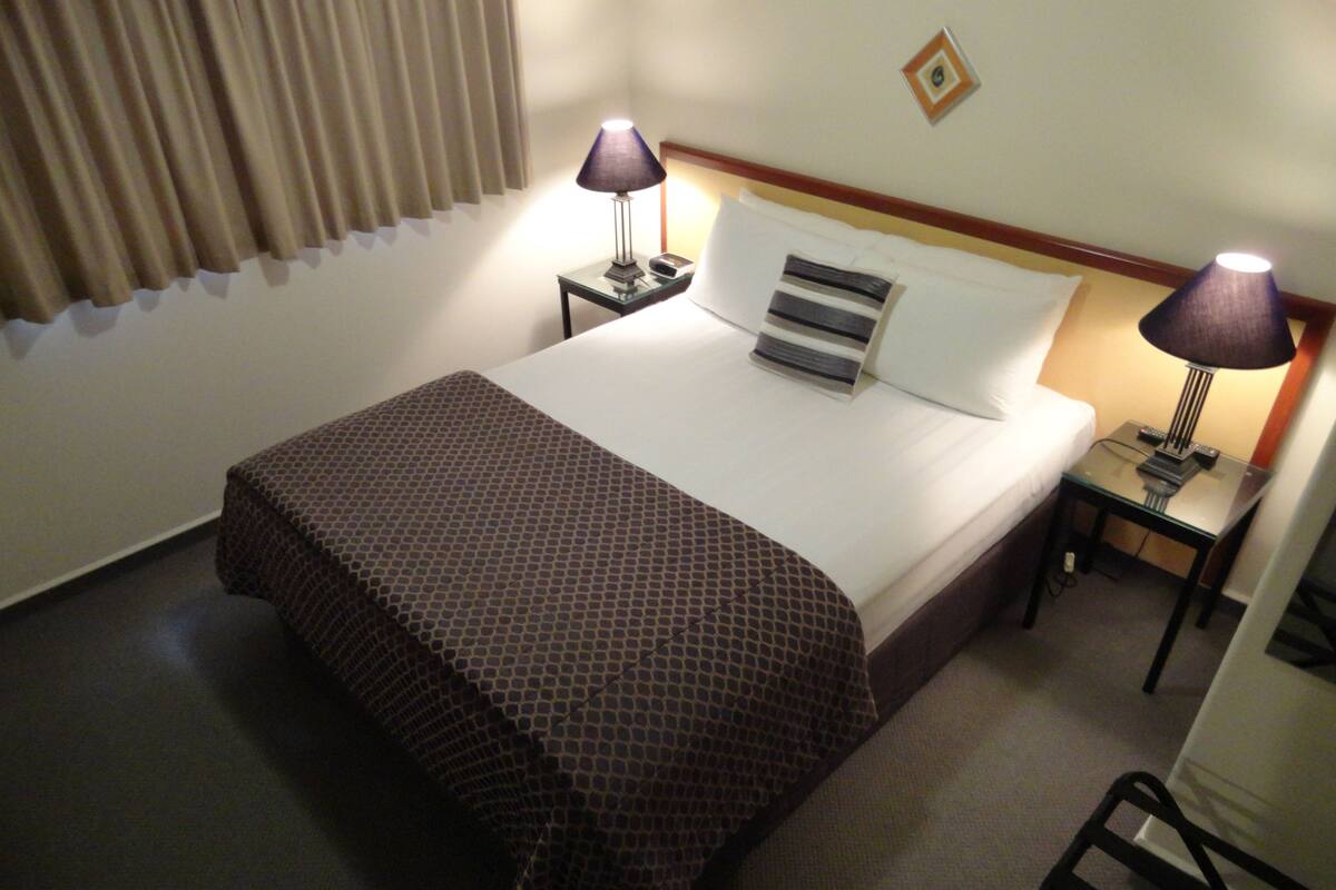 A hotel room with a bed, two bedside tables with lamps, a window with curtains, and a small wall decoration.
