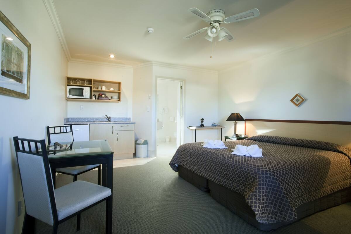 A hotel room with a bed, a desk with a chair, a microwave, a small table with a lamp, and a ceiling fan.