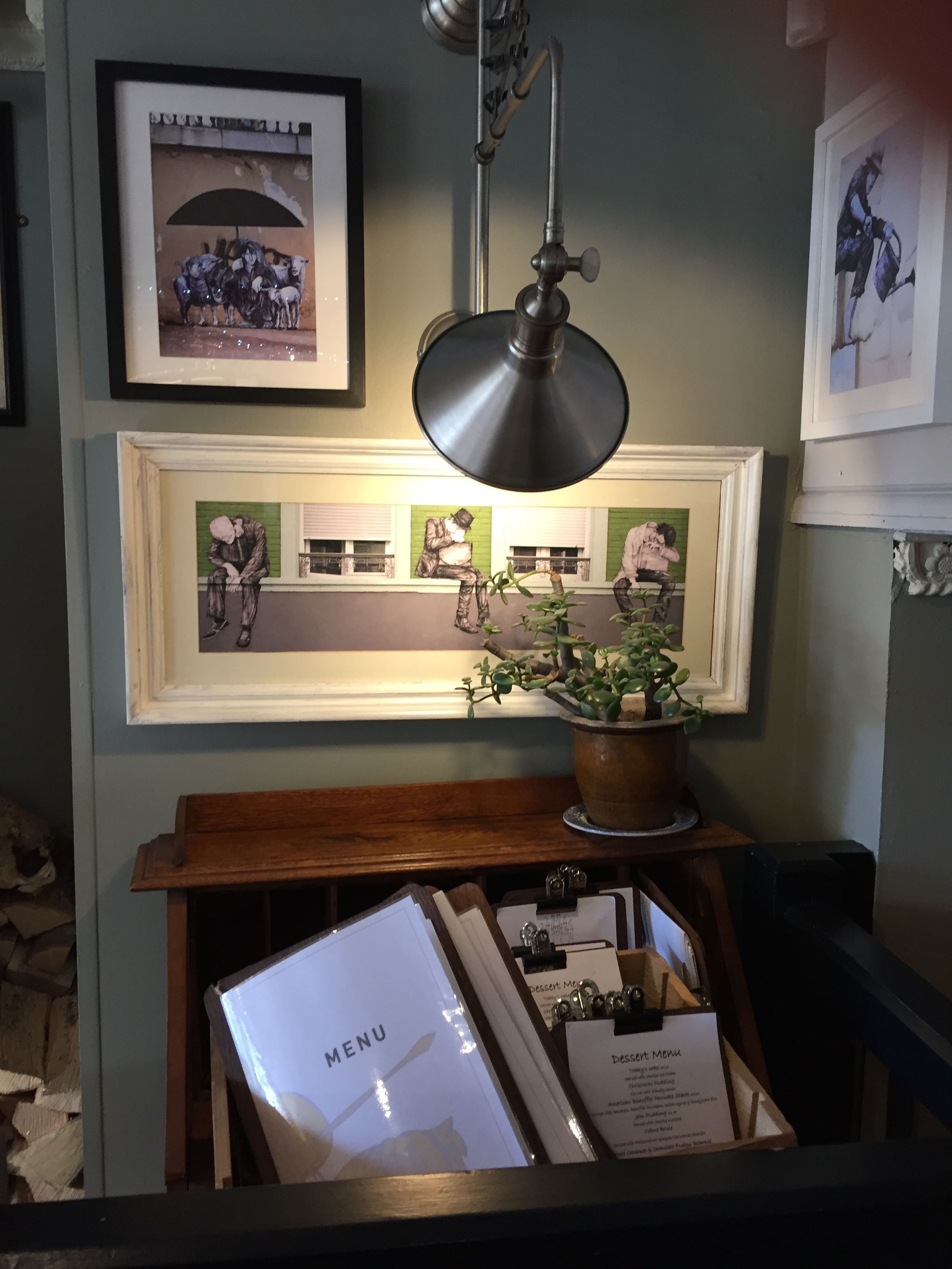 A framed artwork of a street scene, a hanging industrial-style lamp, and a wooden table with a potted plant and menu cards.