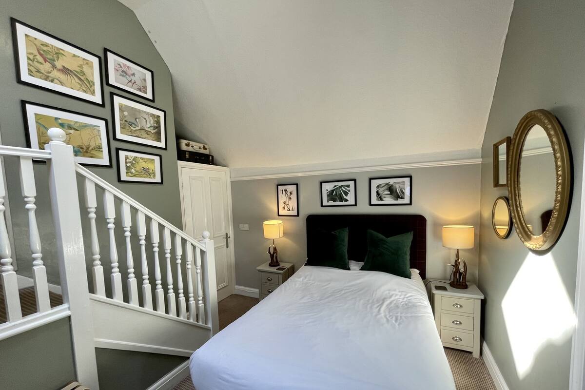 A bedroom with a bed, nightstands, a mirror, a staircase, and framed pictures on the wall.
