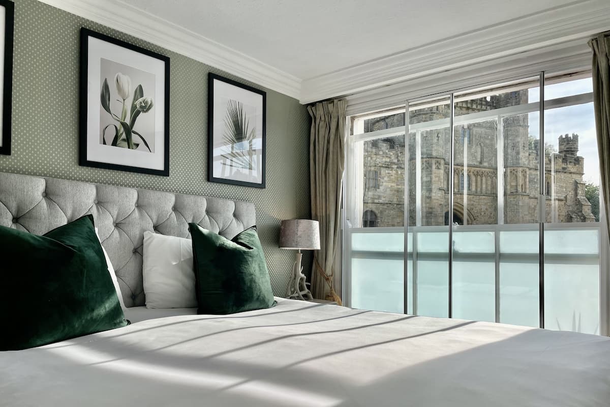 A bedroom with a large bed, a tufted headboard, and a view of historic buildings through a large window.
