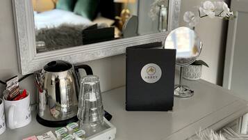 Habitación de hotel con un espejo grande y ornamentado, una mesita de noche con cafetera, un atril con artículos de aseo y un jarrón decorativo con orquídeas.