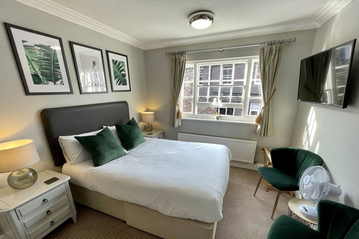A neatly arranged hotel room with a large bed, a bedside table with a lamp, a chair, a small table with a fan, and a window with curtains.