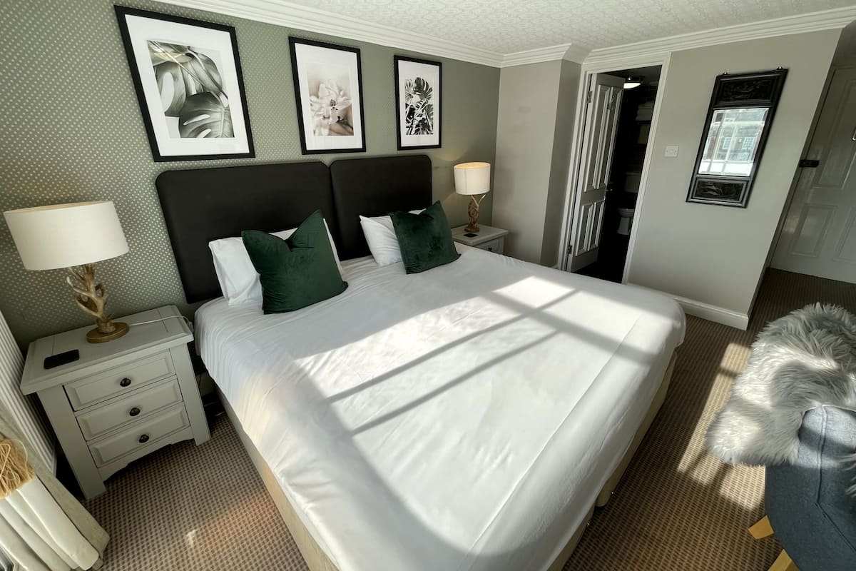 A neatly made bed with white linens and green pillows, a bedside table with a lamp, and framed artwork on the wall.