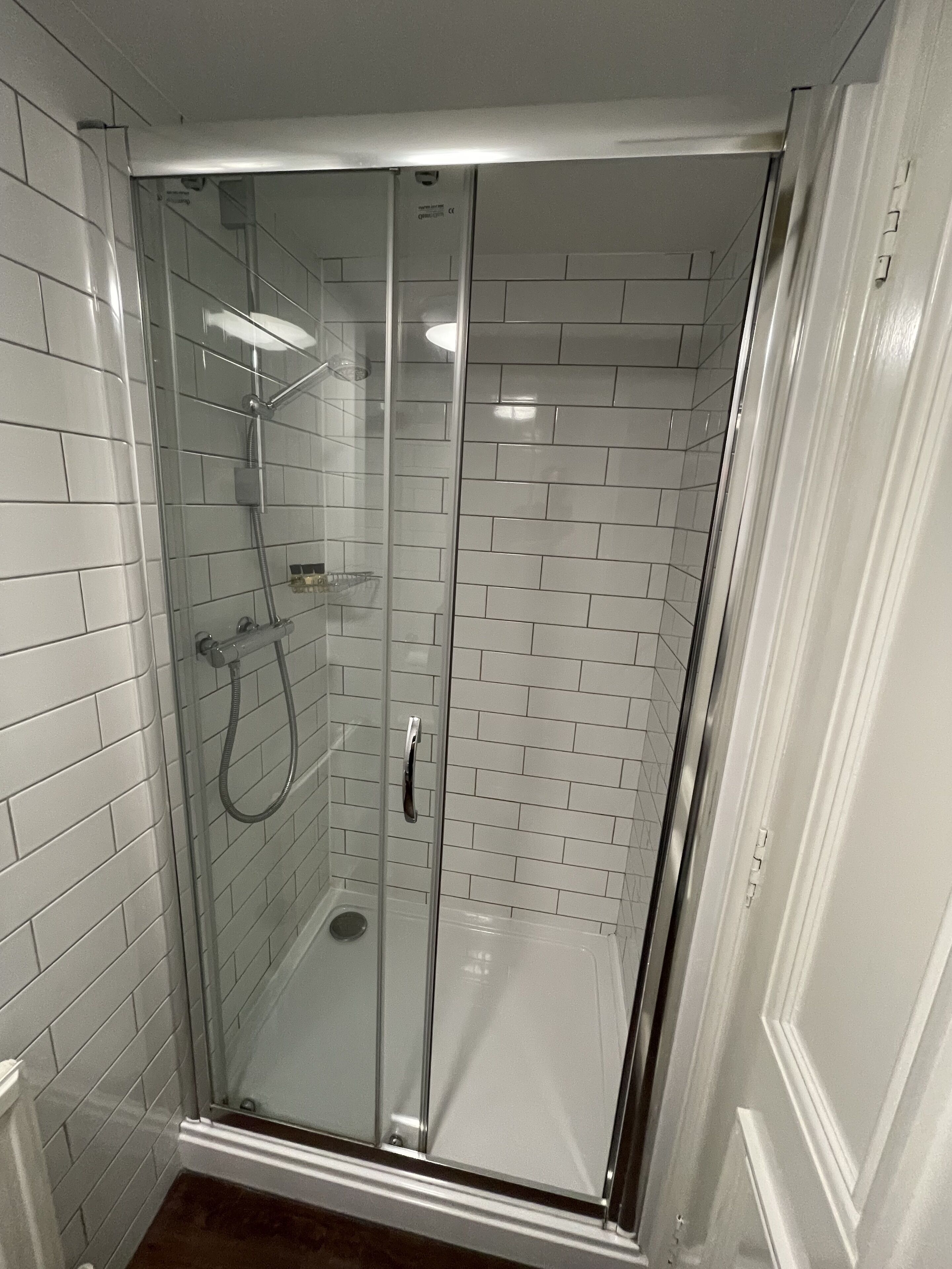 Ducha con mampara de vidrio, paredes de azulejos blancos y puerta blanca.