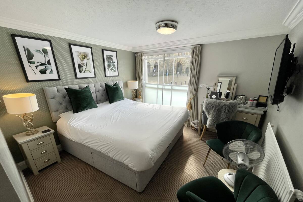 A hotel room with a large bed, a bedside table with a lamp, a chair, and a desk with a computer.