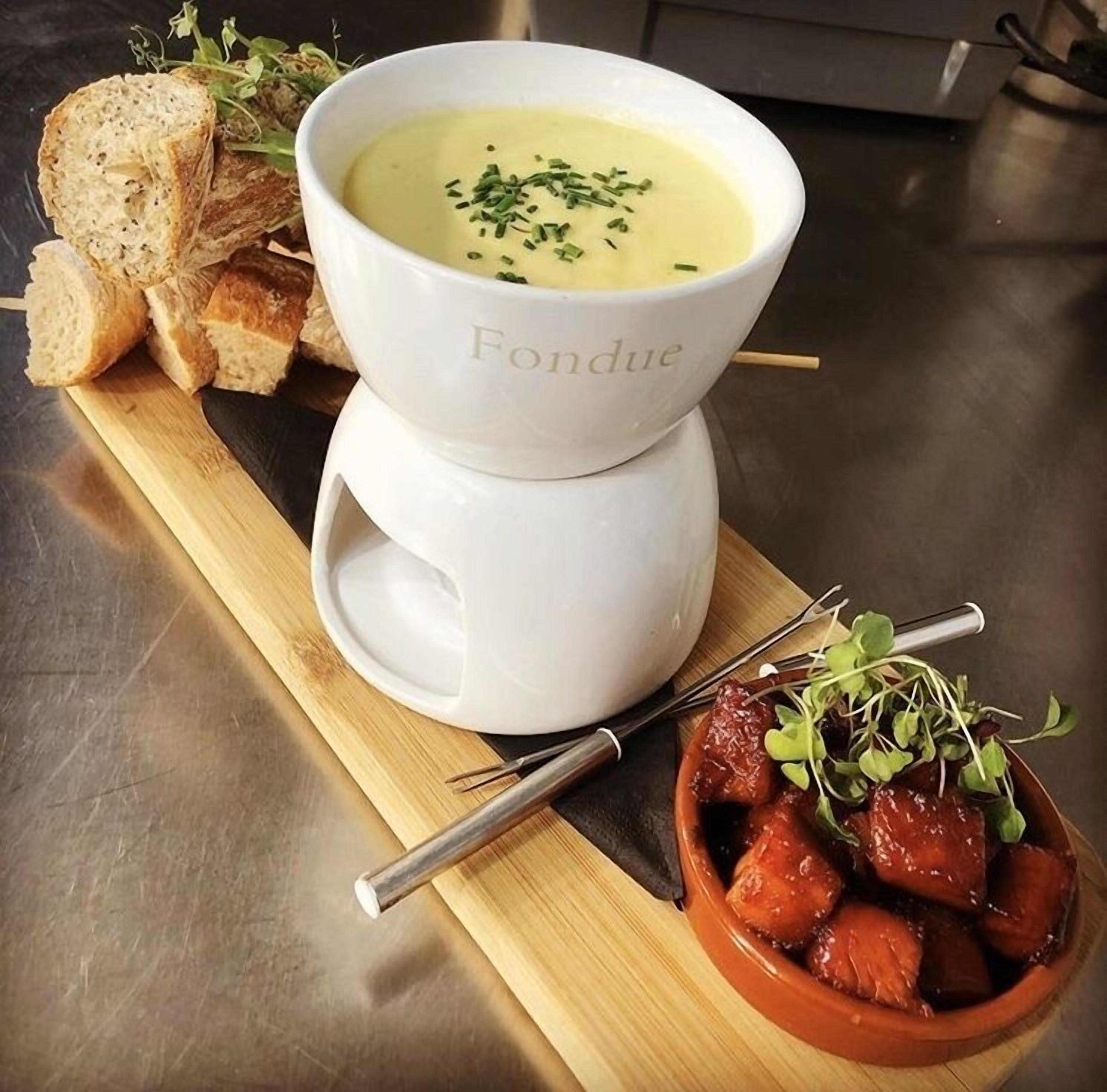 Uma tigela de sopa com uma colher e um prato pequeno de carne, decorado com vegetais, acompanhado de fatias de pão sobre uma tábua de madeira.