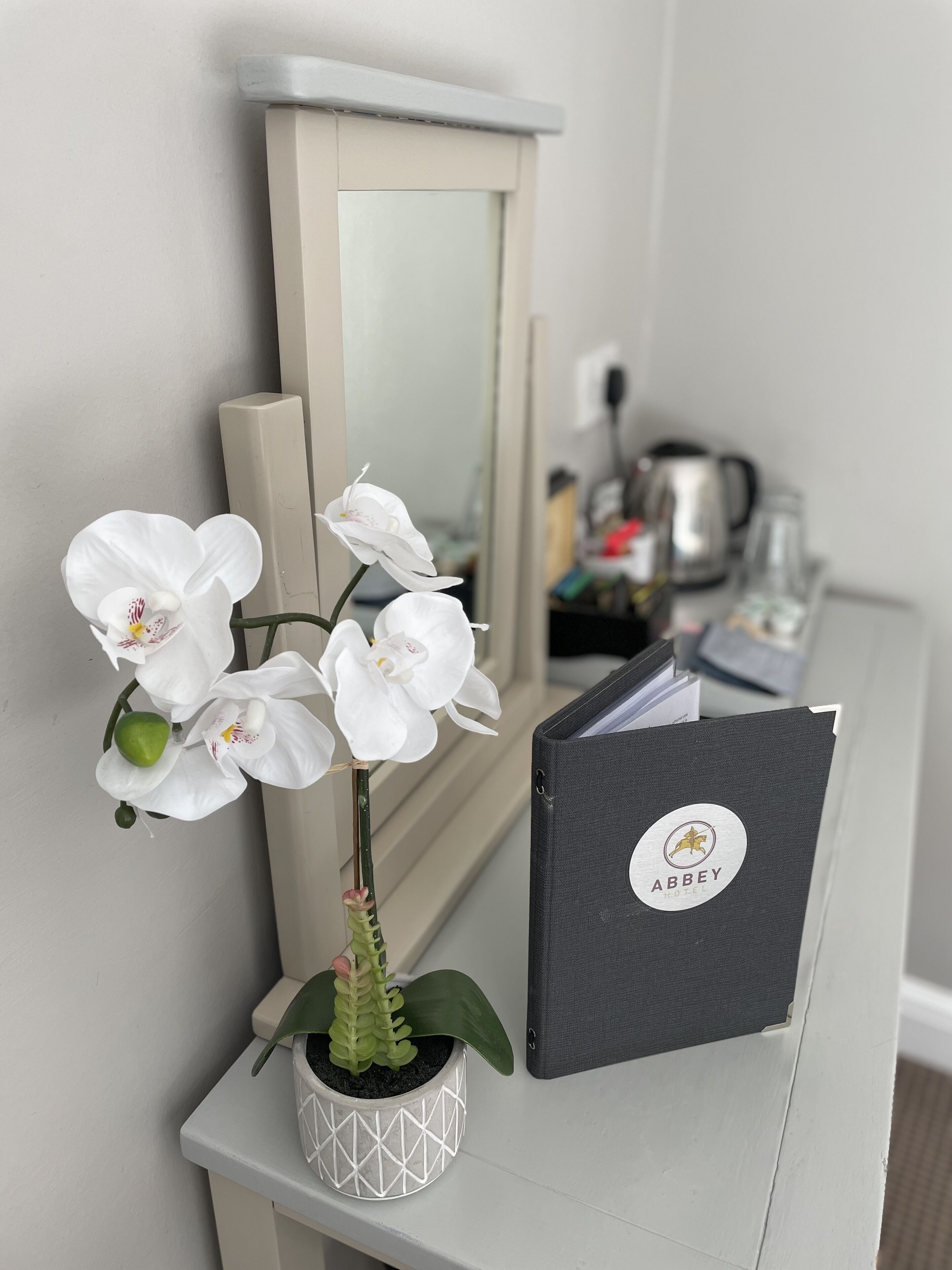 Eine weiße Orchidee in einem weißen Topf, ein schwarzes Buch mit dem 'Abbey Hotel'-Logo und ein Spiegel auf weißer Oberfläche.