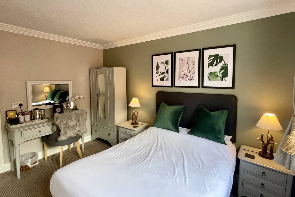 A hotel room with a large bed, a desk with a mirror, a wardrobe, and three framed botanical prints on the wall.