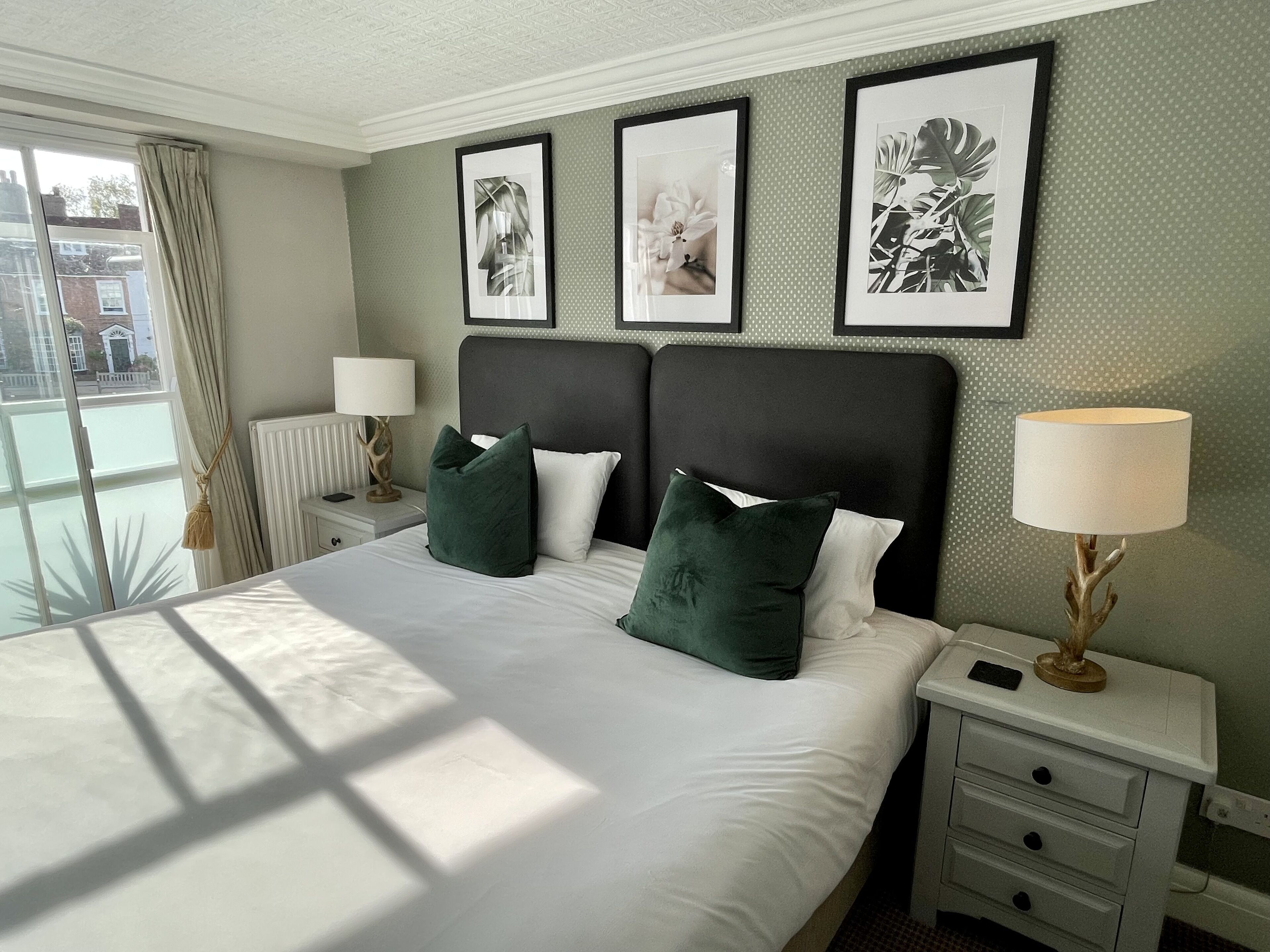 Une chambre d’hôtel avec un grand lit, deux tables de chevet, une fenêtre avec des rideaux et trois tableaux encadrés accrochés au mur.