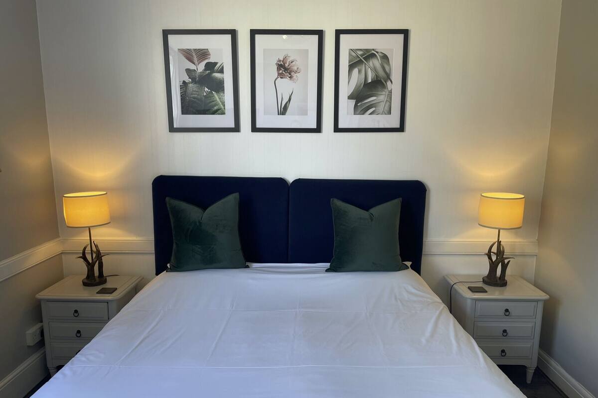 A neatly made bed with a dark headboard, flanked by two bedside tables with lamps and a nightstand drawer. Three framed botanical prints adorn the wall above the bed.