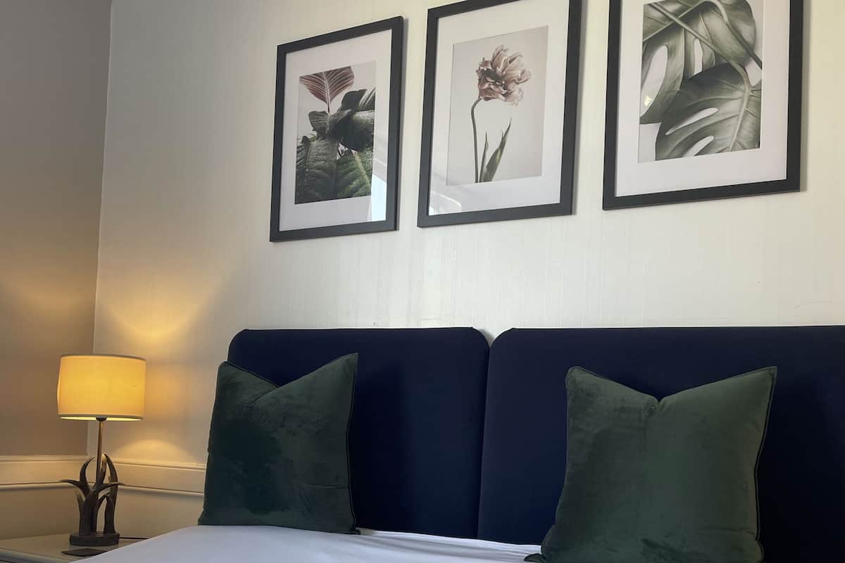 A hotel room with a bed, two green pillows, a nightstand with a lamp, and three framed pictures on the wall.