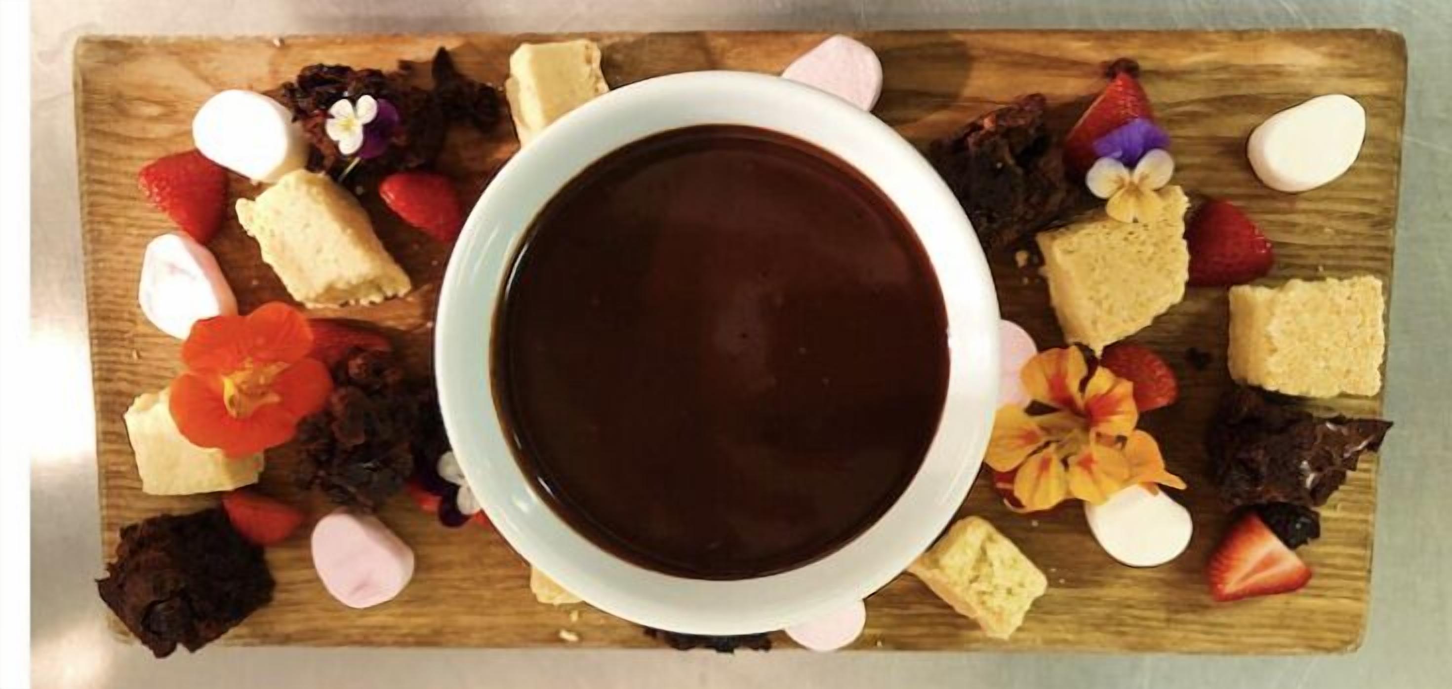 Una taza de chocolate rodeada de varios pasteles y frutas sobre una tabla de madera.