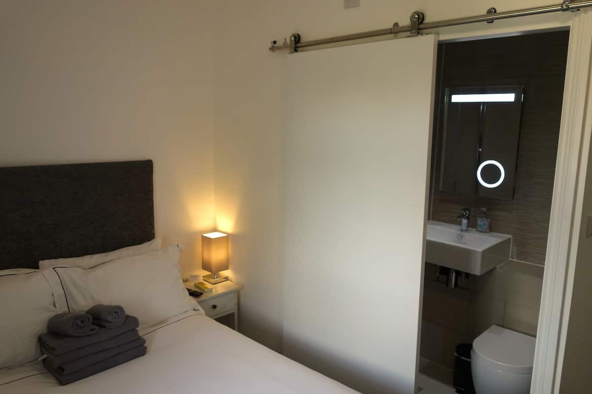A hotel room with a bed, a nightstand with a lamp, a bathroom with a sink and toilet.