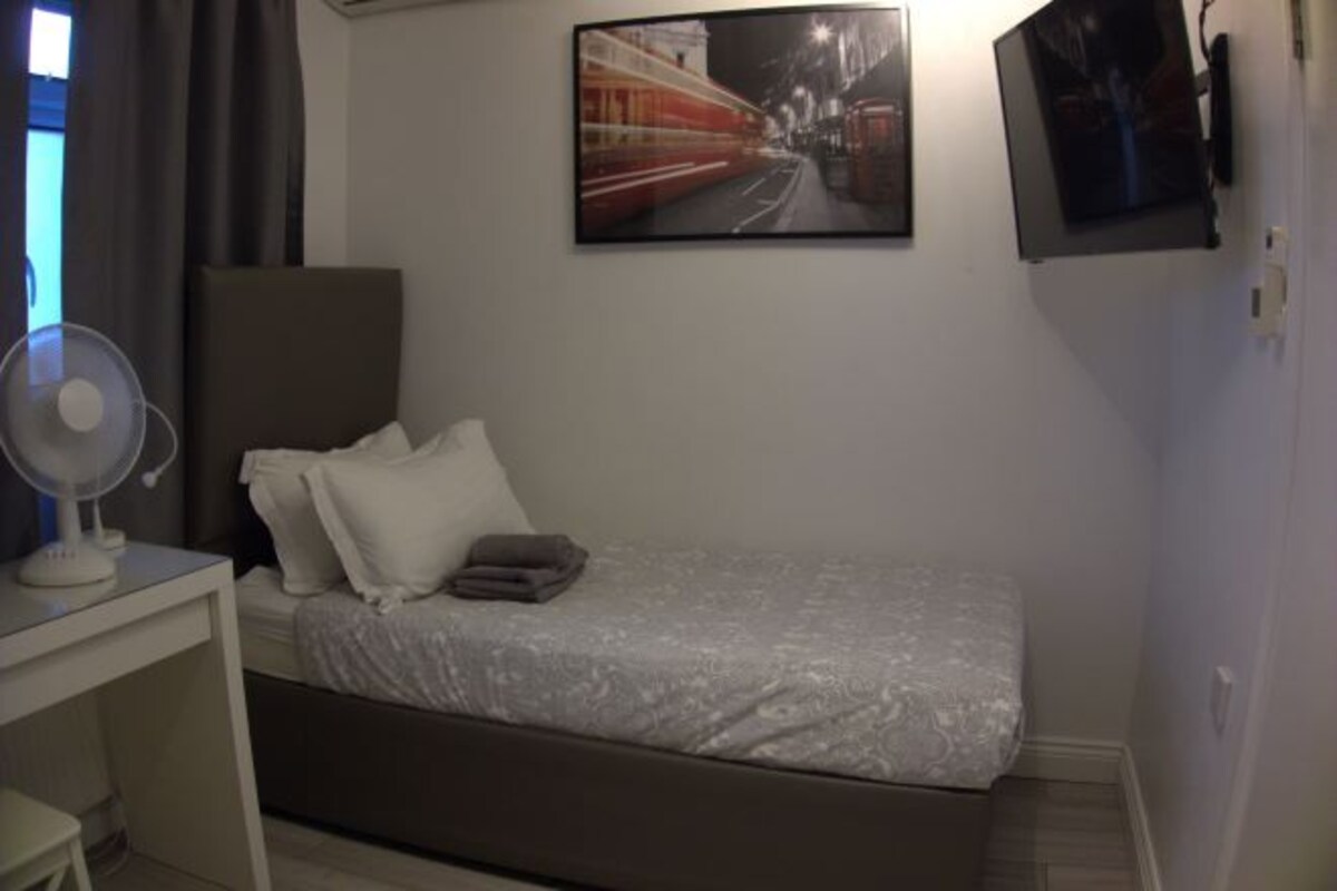 Un dormitorio con cama, escritorio con ventilador, televisor en la pared y un cuadro de un tren.