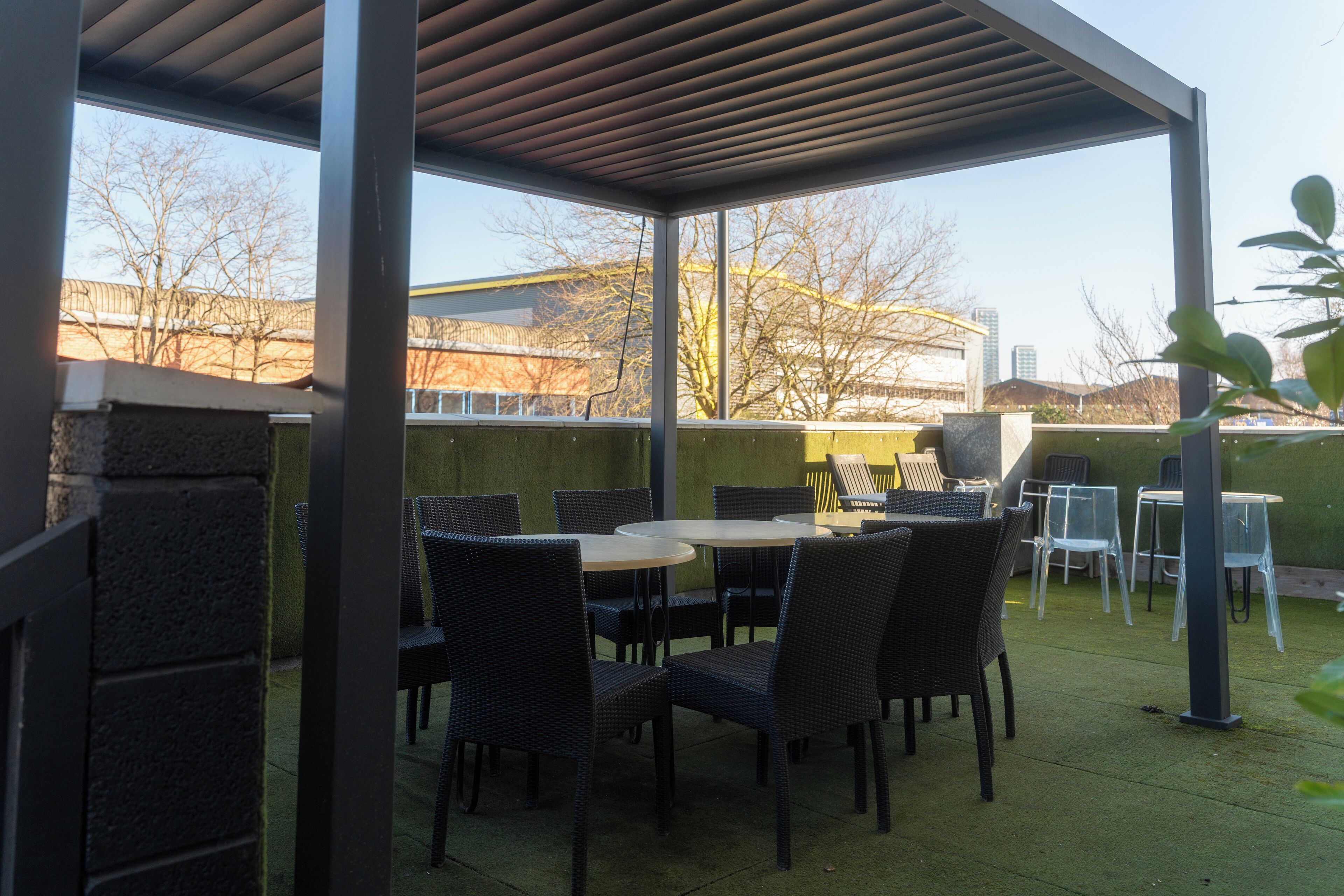 An outdoor patio with wicker chairs and tables, a pergola, and a view of buildings and trees.