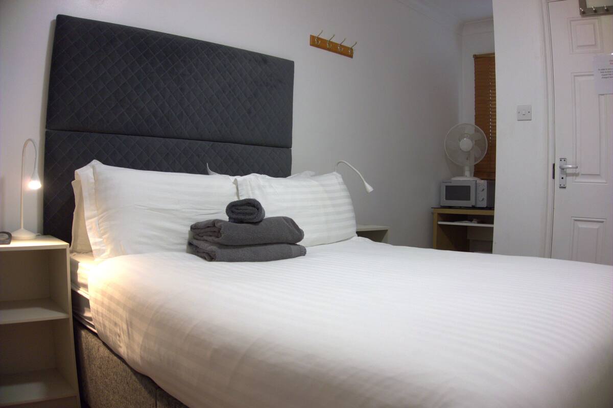 A neatly made bed with white linens and a dark headboard, a bedside table with a lamp, and a small fan on a nightstand.