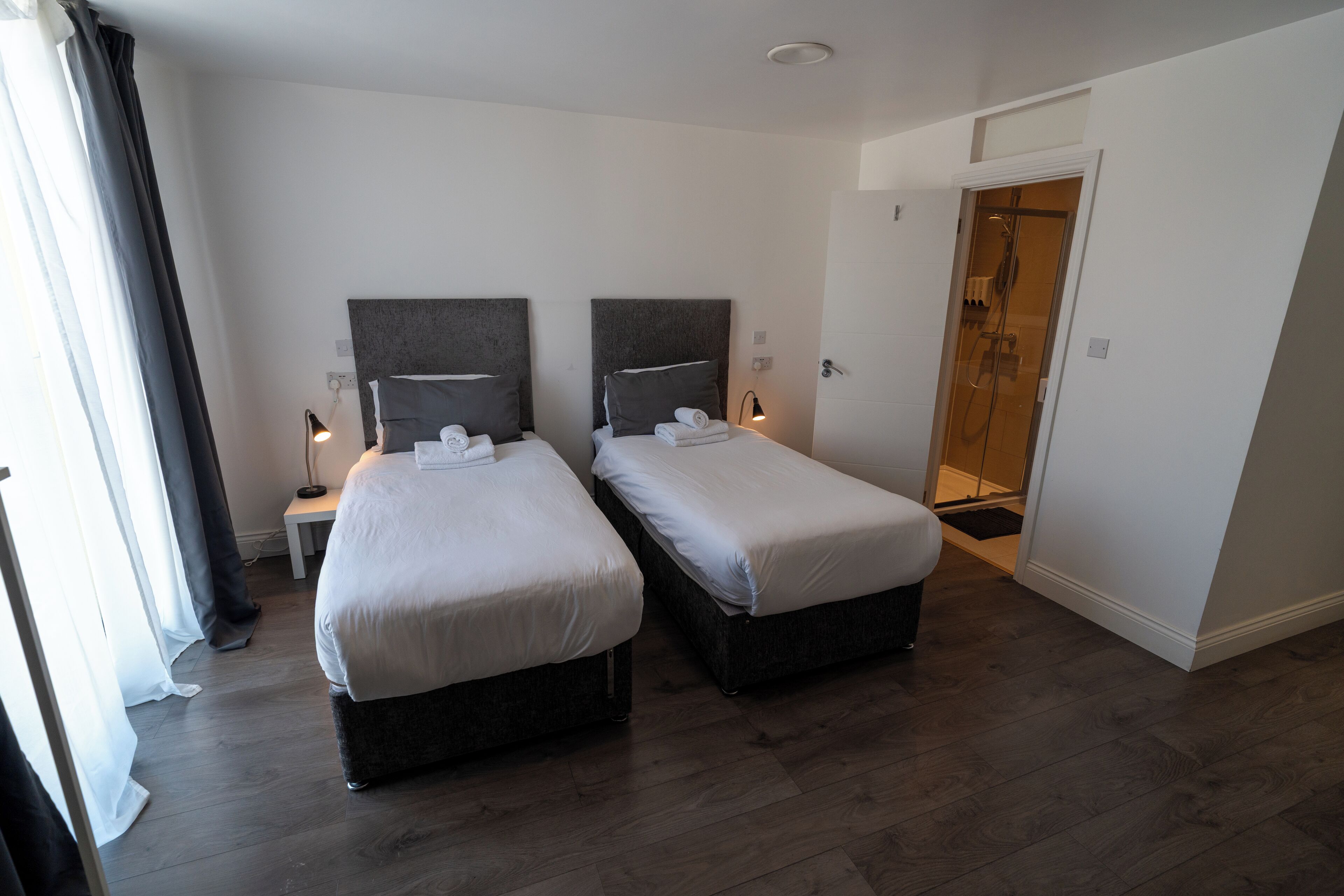 A hotel room with two beds, a shower area, and a small table with a lamp.