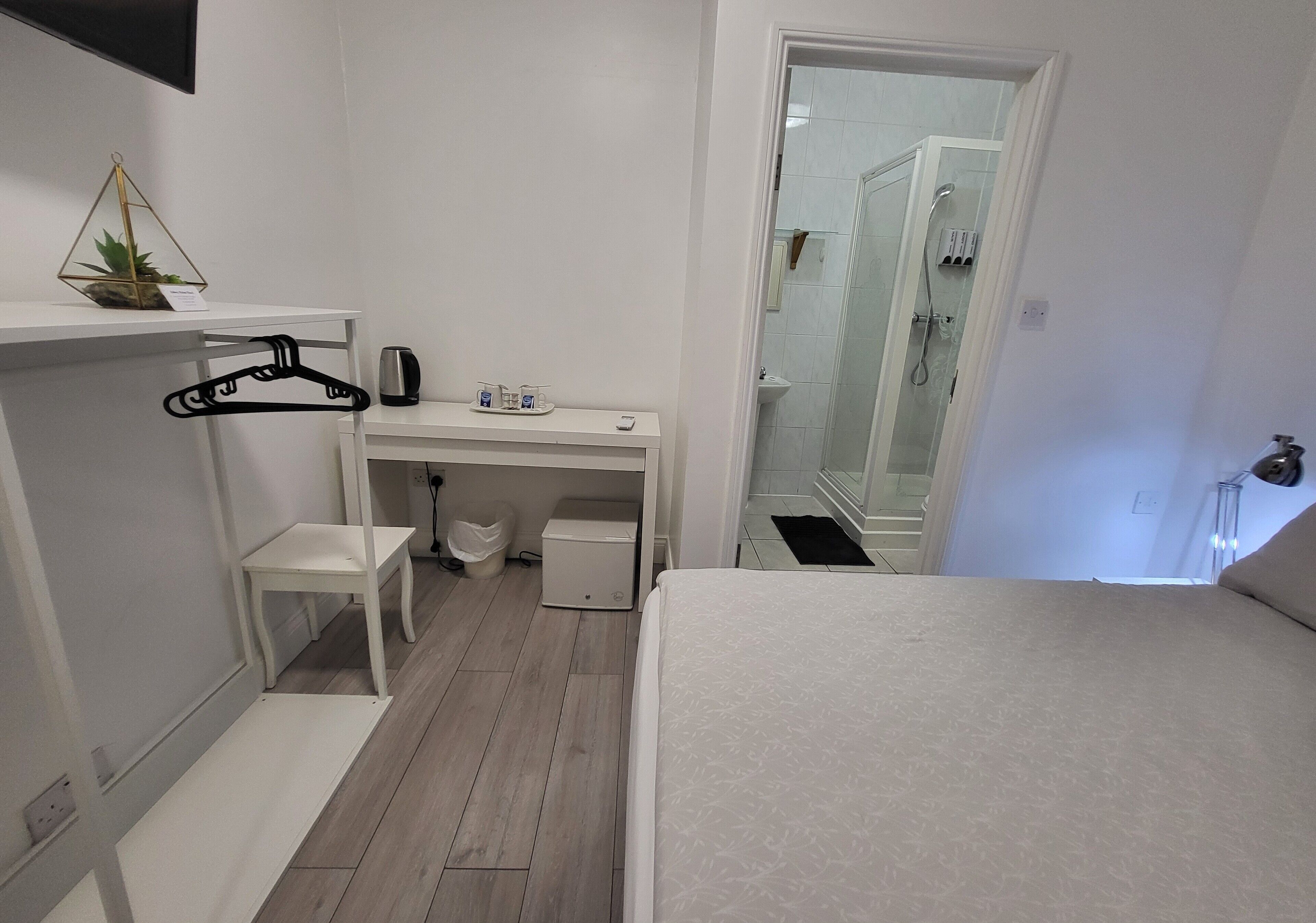 A bedroom with a bed, a desk, a chair, a hanging plant, and a shower area.