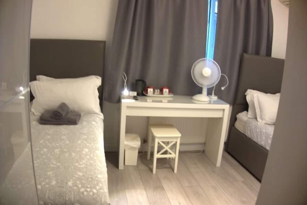 A bedroom with a bed, a desk, a chair, a small stool, and a bedside table with a lamp.