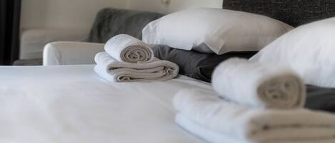 A neatly made bed with white linens and rolled towels, a grey headboard, and a white pillow.