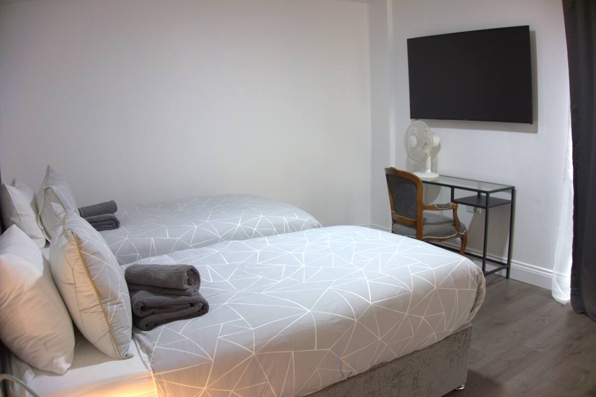 A bedroom with two beds, a desk, a chair, and a flat-screen TV on the wall.