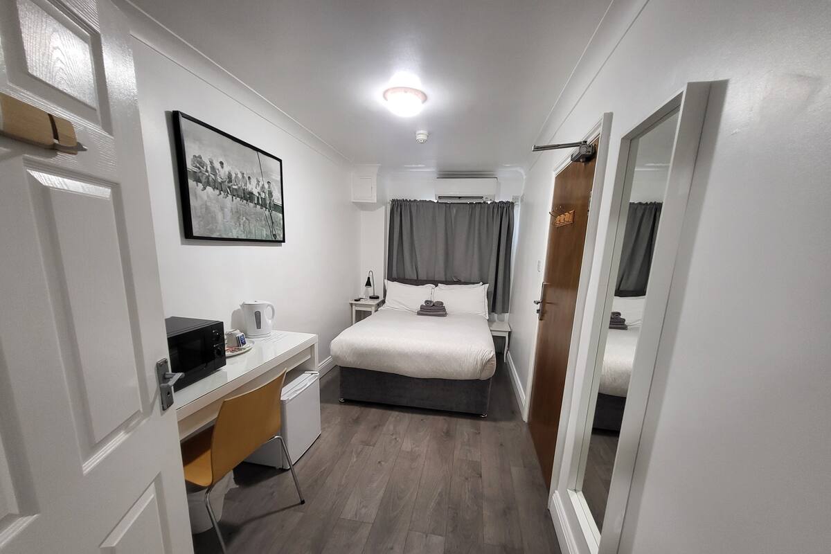 A hotel room with a bed, a desk with a chair, a coffee maker, a framed picture on the wall, and a door with a mirror.