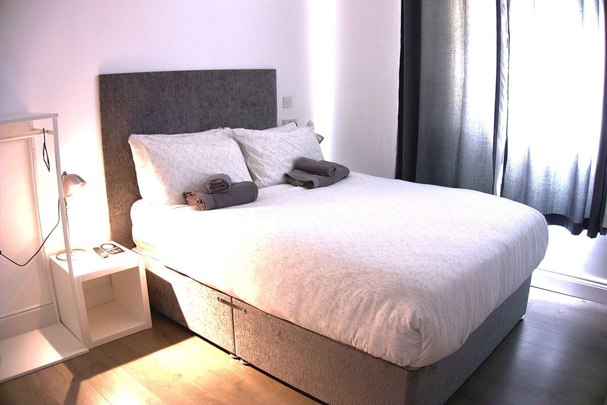 A neatly made bed with pillows and a blanket, a bedside table with a lamp, and a window with curtains.