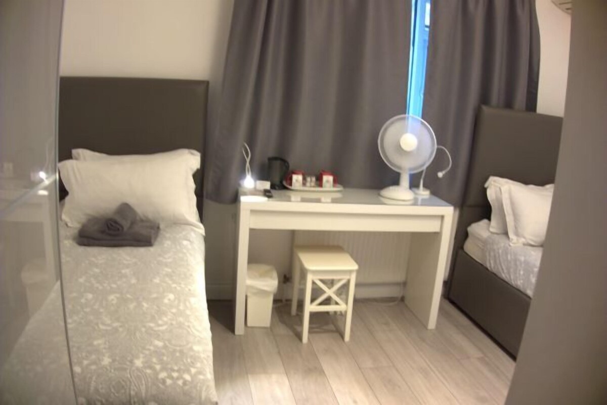 Un dormitorio con cama, escritorio, silla, taburete pequeño y mesita de noche con lámpara.