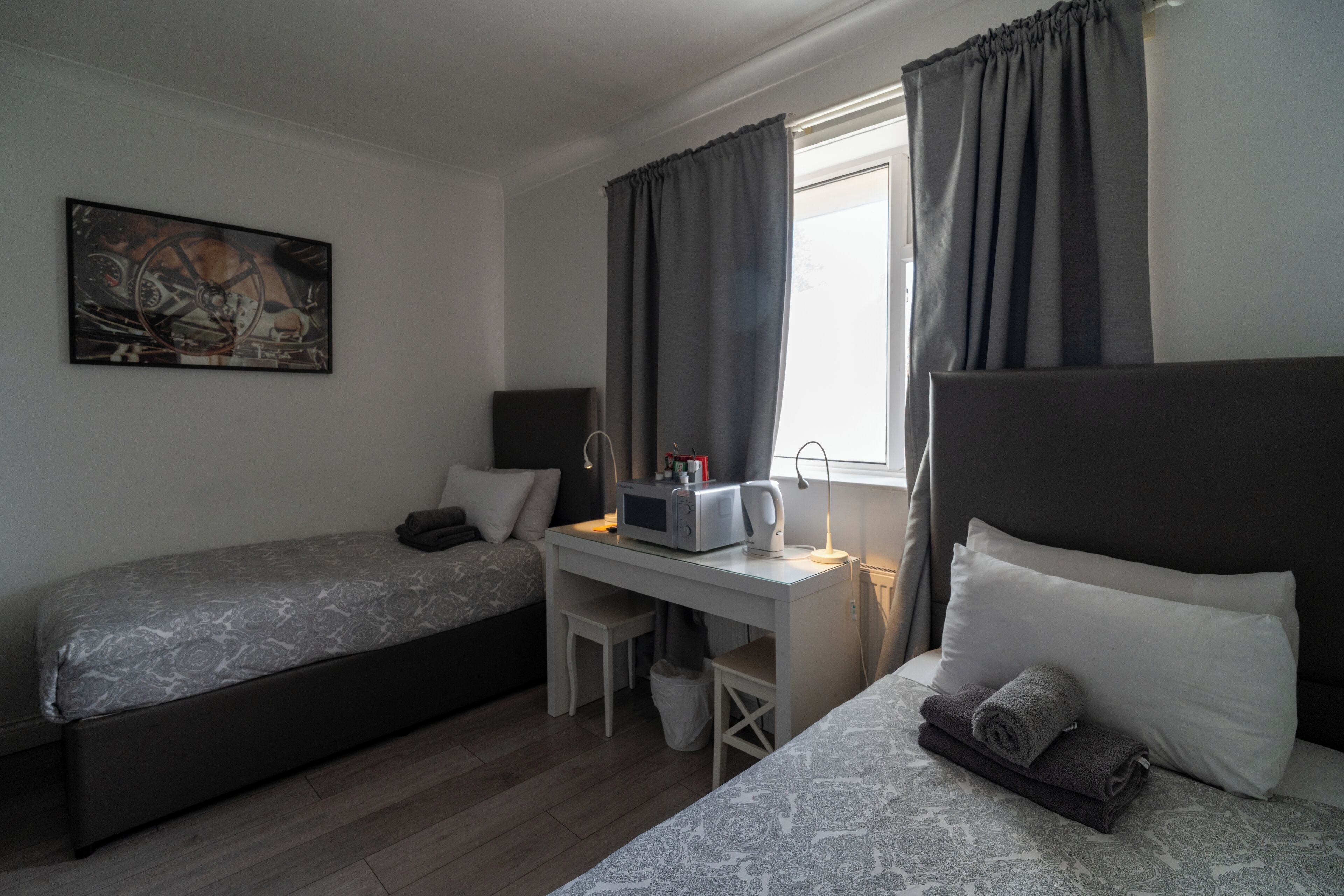 A hotel room with a bed, a desk with a computer, a chair, a window with curtains, and a painting on the wall.