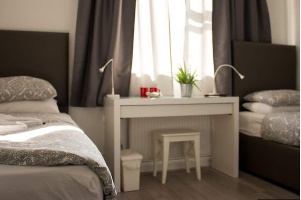 A bedroom with a bed, a desk, a chair, a small stool, a potted plant, and a nightstand.