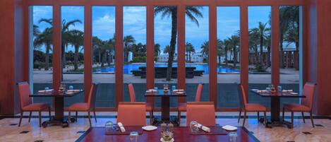 Un comedor con mesas y sillas, ventanas amplias que dan a palmeras y una piscina.