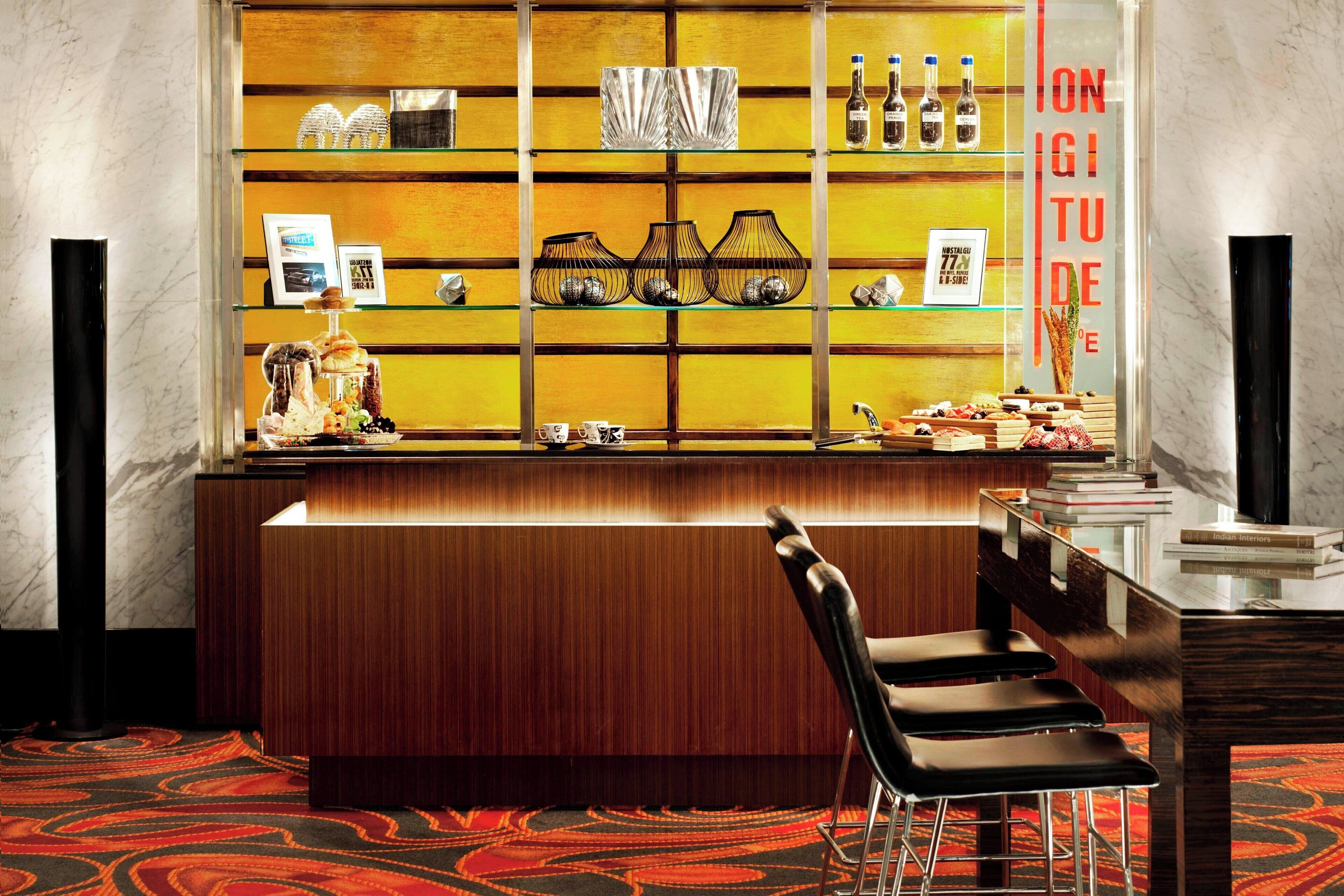 Una zona de bar con mostrador de madera, taburetes negros y estantes con diversos artículos.