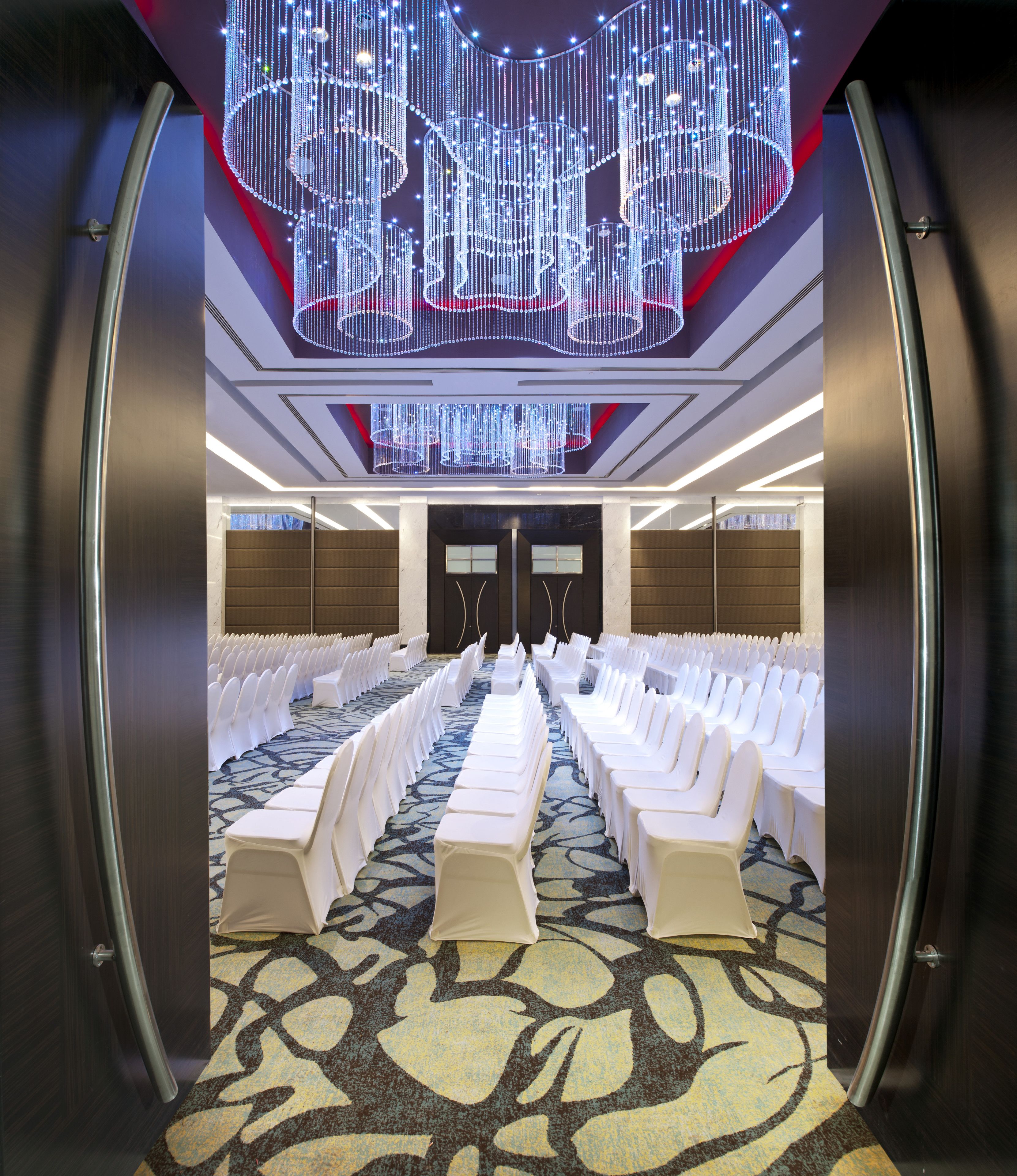 Une salle de conférence avec des rangées de chaises blanches, une moquette à motifs et un lustre moderne.