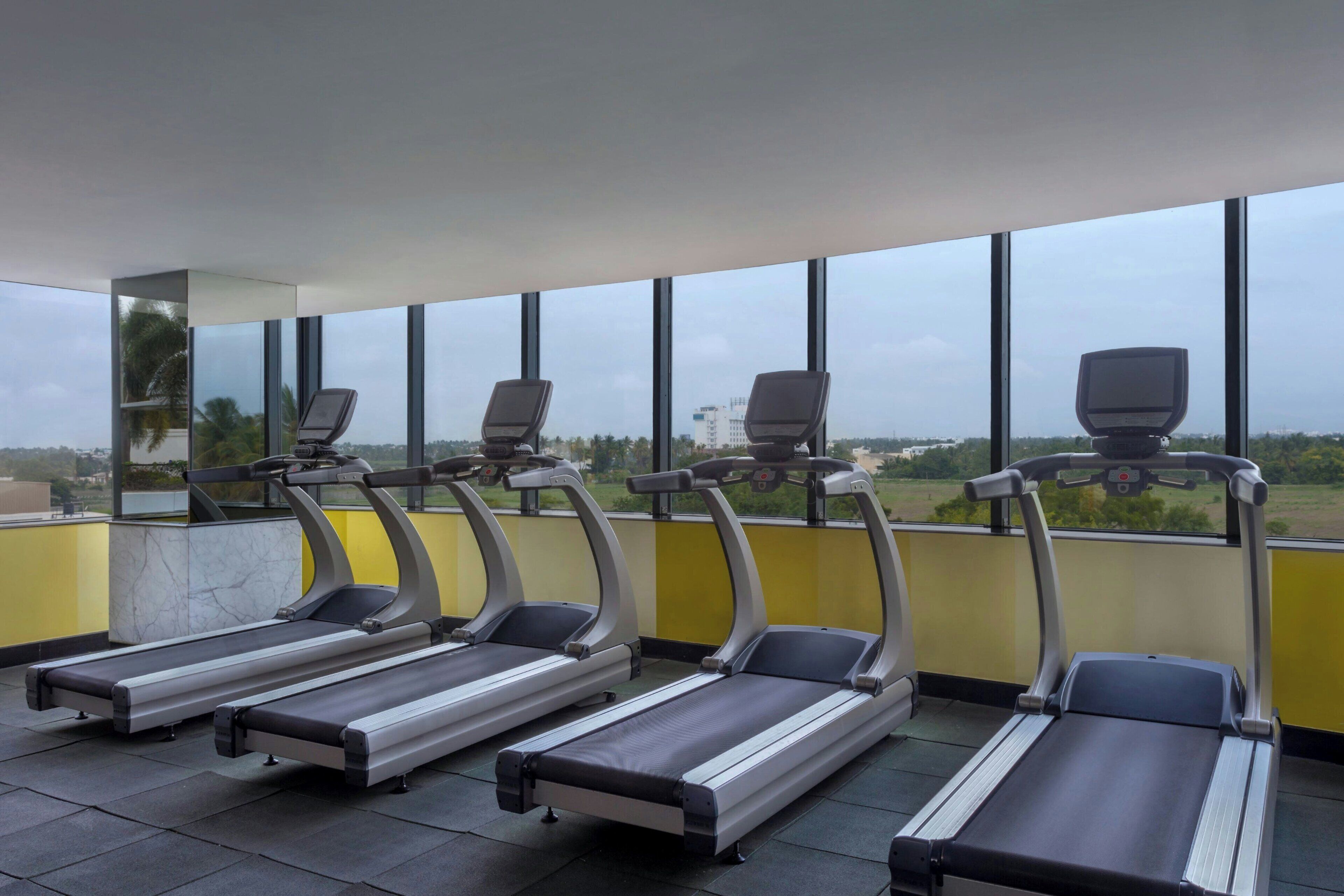 A gym with four treadmills aligned against a wall with large windows, offering a view of the cityscape.