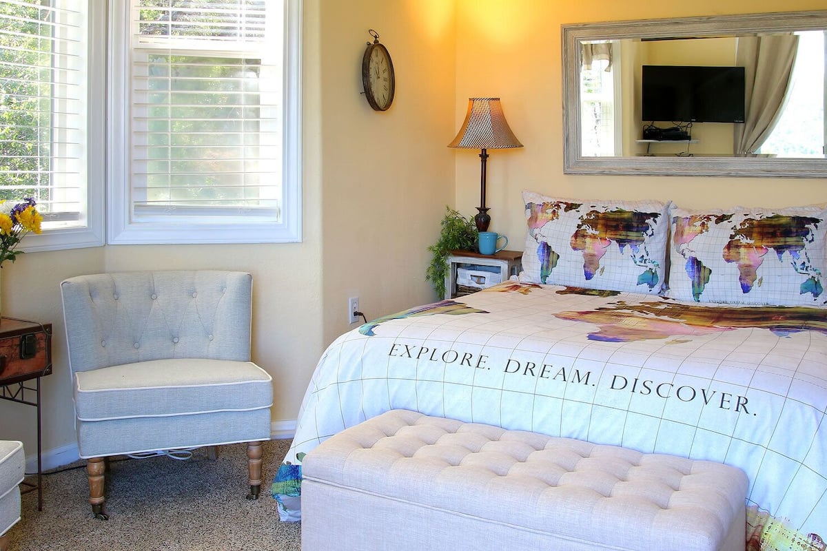 A bedroom with a bed featuring a world map design, a chair, a bedside table with a lamp, and a window with blinds.