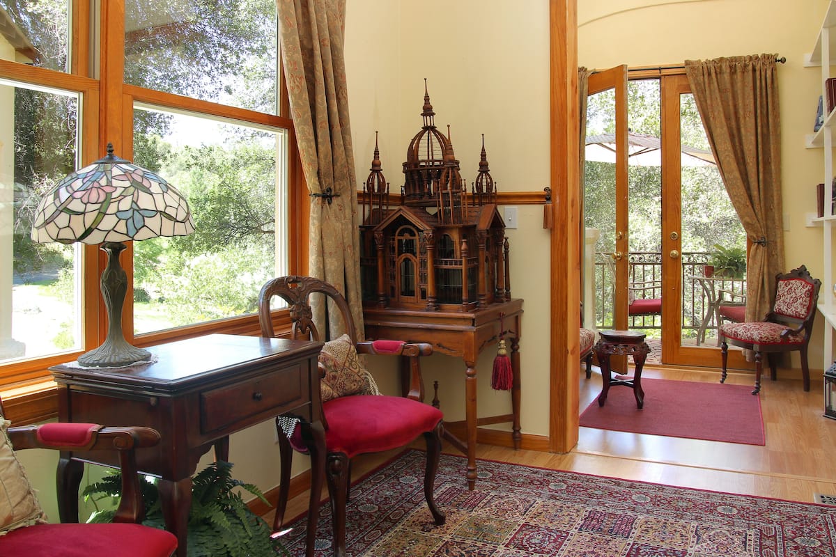 A room with a wooden desk, a red chair, a stained glass lamp, and a large window with curtains.