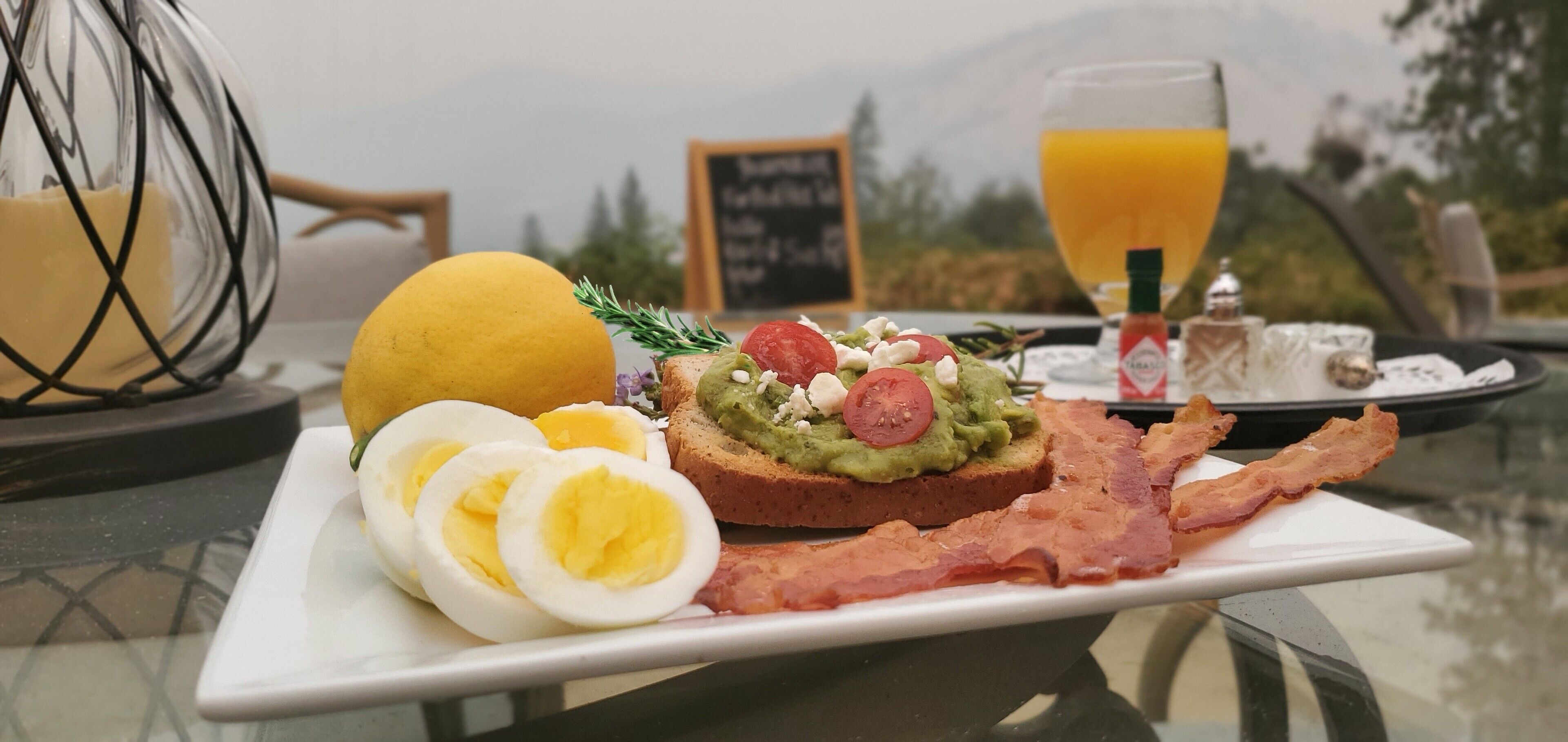 A breakfast plate with avocado toast, bacon, and eggs, a glass of orange juice, and a candle holder on a table with a view of mountains.