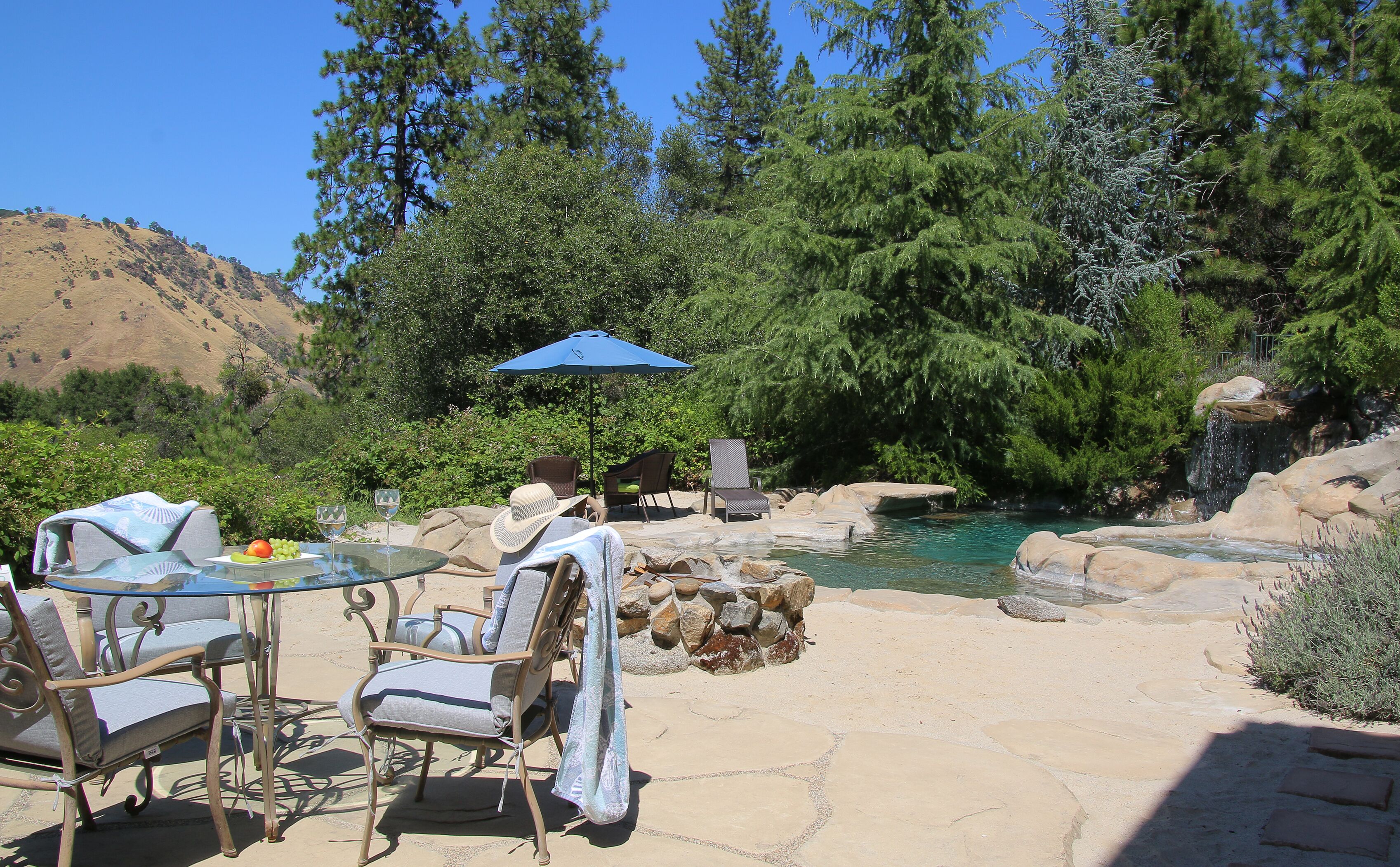 Un patio exterior con una mesa, sillas y un paraguas azul. Hay una piscina natural rodeada de rocas y árboles.